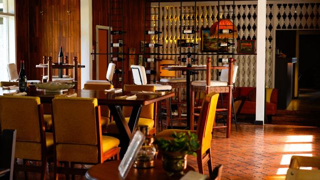 Salón con mesas de madera, sillas tapizadas en amarillo y una pared con estantes de botellas de vino.