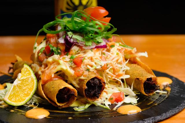Tacos enrollados fritos cubiertos de repollo, tomate y salsa, con una rodaja de naranja, sobre piedra negra.