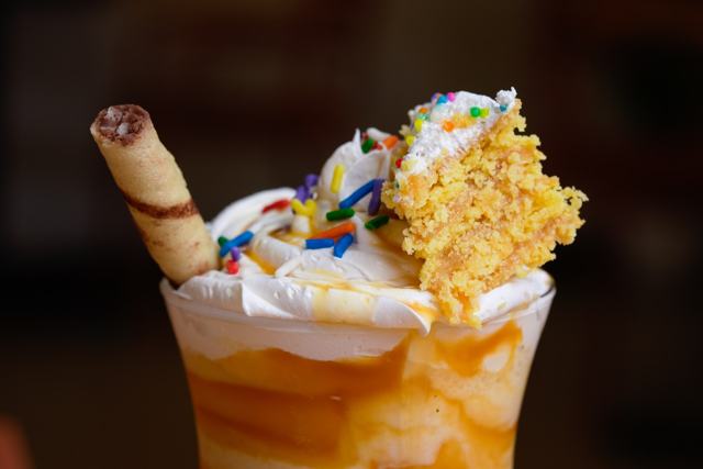 Milkshake de caramelo en vaso de vidrio con crema batida, confites de colores, un trozo de queque y un barquillo.