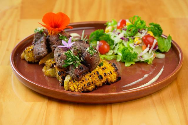 Trozos de carne a la parrilla con elote asado y ensalada, decorados con flores, en plato de barro.